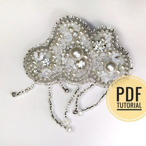May include: A white beaded cloud brooch with pearl accents and dangling beaded strands. PDF TUTORIAL is written on a yellow circle in the lower right corner of the image.