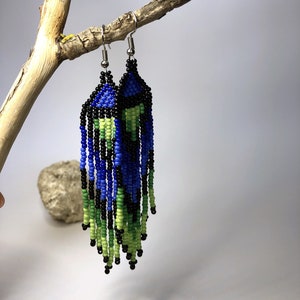 May include: A pair of beaded earrings with a blue, green, and black color scheme. The earrings are hanging from a branch.