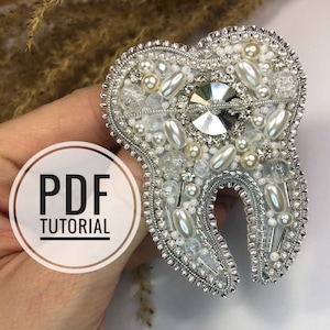May include: A silver tooth-shaped brooch with white beads and a clear rhinestone in the centre. The brooch is on a hand and the text "PDF TUTORIAL" is in a circle on the hand.