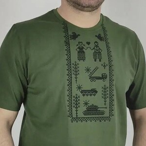 May include: A green t-shirt with a black embroidered design featuring a couple, a truck, and a tank. The design is framed by a border of black stitching.