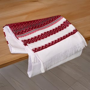 May include: White embroidered table runner with red and black geometric patterns and white fringe.