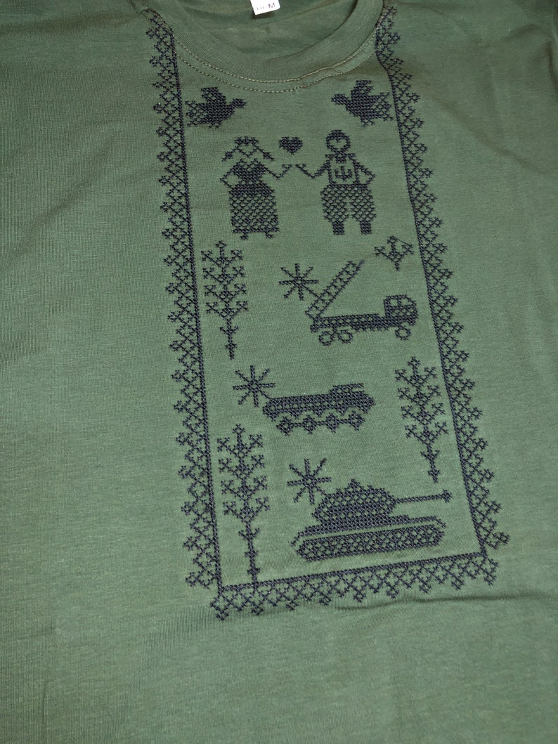 May include: Green t-shirt with a black embroidered design. The design features a couple, trees, a truck, a tank, and a snowflake.