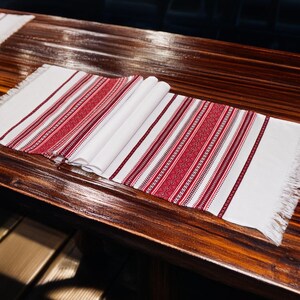May include: A white and red striped table runner with fringed edges is displayed on a dark wooden table. The runner features a repeating pattern of red stripes and decorative stitching.