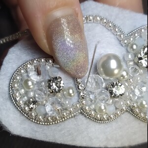 K&ouml;nnte beinhalten: Nahaufnahme einer herzf&ouml;rmigen Brosche aus wei&szlig;em Filz mit silbernen Perlen, Perlen und Strasssteinen. Die Brosche wird von einem Finger mit glitzerndem hellrosa Nagellack gehalten.