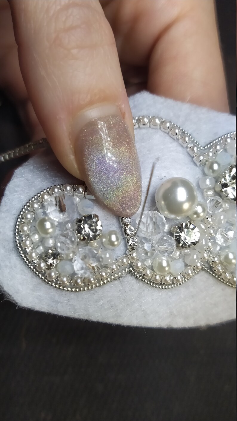 K&ouml;nnte beinhalten: Nahaufnahme einer herzf&ouml;rmigen Brosche aus wei&szlig;em Filz mit silbernen Perlen, Perlen und Strasssteinen. Die Brosche wird von einem Finger mit glitzerndem hellrosa Nagellack gehalten.