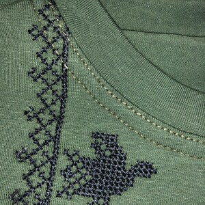 May include: A green t-shirt with black embroidered designs. The embroidery features a geometric pattern and a bird-like figure.