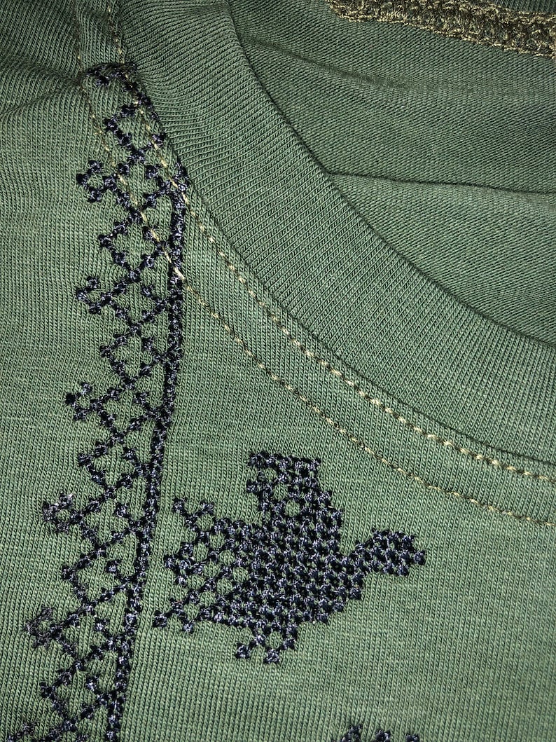 May include: A green t-shirt with black embroidered designs. The embroidery features a geometric pattern and a bird-like figure.