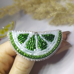 Op de afbeelding: Een broche in de vorm van een limoenwig met groene en witte kralen. De broche is gemaakt van kleine, ronde kralen die aan elkaar zijn genaaid om de vorm van een limoenwig te creëren. De broche is een geweldige manier om een ​​toets van whimsy toe te voegen aan elke outfit.