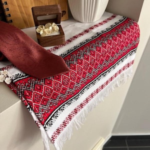 Può includere: Un runner da tavolo bianco con un motivo geometrico rosso e nero e frange bianche. Una cravatta bordeaux è appoggiata sul runner.