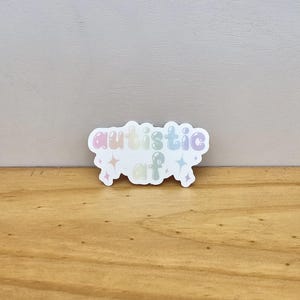 May include: A white sticker with the text "autistic af" in pastel colors. The sticker has a cloud-like shape with star accents. The sticker is on a wooden surface.
