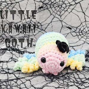 May include: A rainbow-colored crocheted spider with a black bow, sitting on a black spiderweb background. The text "LITTLE KAWAII GOTH" is visible in the background.