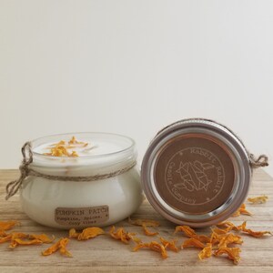 Puede incluir: Una vela en tarro de cristal transparente con cera blanca, adornada con pétalos de naranja secos. El tarro está envuelto con hilo y tiene una etiqueta que dice "Pumpkin Patch". La tapa está a un lado, revelando el logo de Rabbit Candle Company.