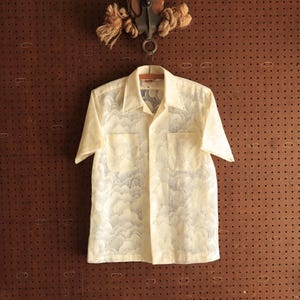 May include: Cream-colored short-sleeved shirt with a cloud-like pattern. The shirt has two chest pockets and a classic collar. It hangs from a metal hook against a brown pegboard background, with a rope and pulley detail at the top.