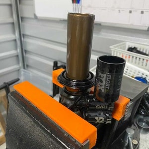 May include: A bicycle suspension fork being worked on, clamped in a vise. The fork has a brown tube, a black tube with "FOX FLOAT" printed on it, and orange protective jaws on the vise.