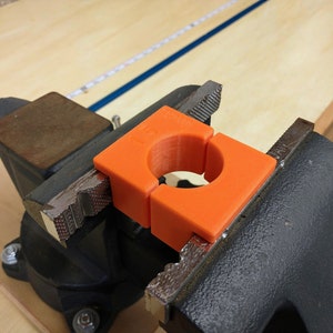 May include: An orange, 3D-printed clamp is held in a black metal vise. The clamp has a circular opening and is designed to protect the object being held. The vise is on a wooden surface.