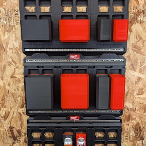 May include: Three black Milwaukee Packout wall plates mounted vertically, featuring various modular storage attachments in black and red. The system includes rectangular bins and holders, with one holder containing a silver and red soda can.