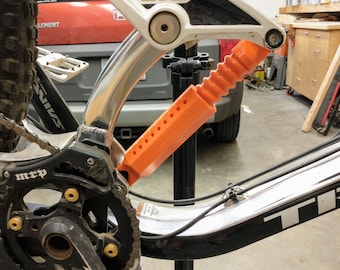 May include: Close-up of a mountain bike's rear derailleur and chainring. The bike is silver and black with a black chain and a black chainring. The chainring has the word "MRP" printed on it. The bike is in a garage with a red car in the background.