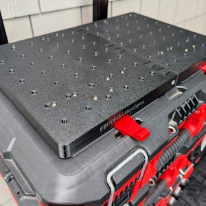 Puede incluir: Una superficie de trabajo rectangular negra con una cuadrícula de orificios circulares, que presenta el texto "@NoksDesigns" grabado en el lateral. El tablero está montado sobre una caja de herramientas modular negra y roja con cierres rojos.