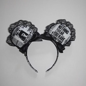 May include: Black and white headband with two circular ear-shaped accents. Each accent features a newspaper-style print with text and a black lace trim. Black bows are attached at the base of each accent.