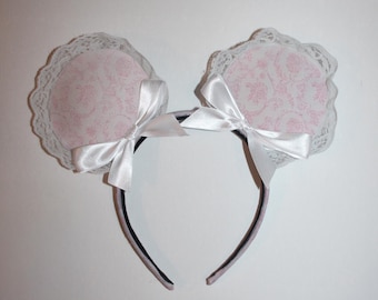 Puede incluir: Una diadema con dos decoraciones circulares en forma de oreja. Cada oreja presenta un estampado floral rosa, un ribete de encaje blanco y un lazo de satén blanco. La diadema es negra y rosa.