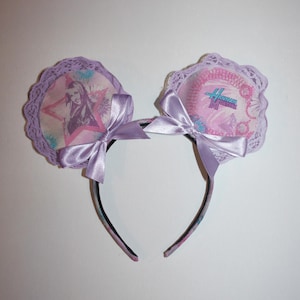 May include: A headband with two circular decorations featuring images and text. The decorations are edged with lilac lace and adorned with lilac bows. One decoration has a star and a person's image, the other has the text "Hannah Montana".