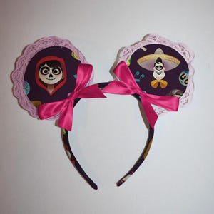 May include: A headband with two circular ear-shaped decorations featuring characters from the film Coco. The ears are purple with pink lace trim and a bright pink bow. The headband is dark with a colourful pattern.