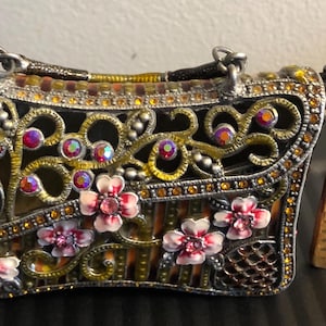 May include: Ornate, vintage-style handbag with a silver frame, embellished with gold filigree, iridescent rhinestones, and pink floral accents. The bag has a small handle and a chain strap. A wooden ruler is next to the bag.