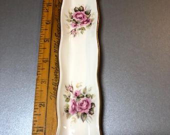 May include: A white porcelain spoon rest with a gold trim and a floral rose design. The spoon rest is approximately 7 inches long and features two clusters of pink roses with green leaves.