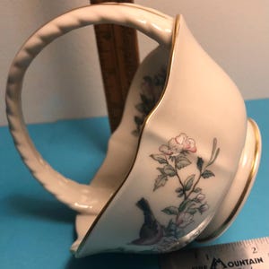 May include: Cream-colored porcelain basket with a curved handle and gold trim. The basket features a floral and bird design in soft pinks, greens, and blues. The handle has a twisted rope design.