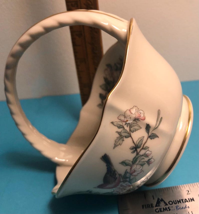 May include: Cream-colored porcelain basket with a curved handle and gold trim. The basket features a floral and bird design in soft pinks, greens, and blues. The handle has a twisted rope design.