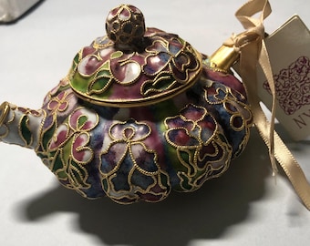May include: A decorative, miniature teapot with a colorful floral design. The teapot features a gold-colored trim and handle, with a lid and a small knob on top. The colors include shades of purple, green, and white. A tag with the letters "NYCC" is attached.