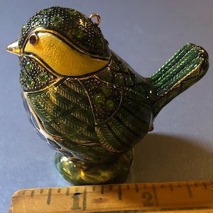 May include: A decorative bird figurine with a yellow breast, green and gold body, and a small loop for hanging. The bird is embellished with green gemstones and has a gold beak. The figurine is approximately 2 inches long.