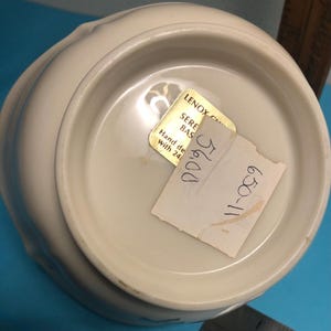 May include: Off-white ceramic bowl with a scalloped rim. The bottom of the bowl has a gold sticker that reads "LENOX SERENE BAS" and a handwritten price tag. The bowl is hand decorated with 24K gold.
