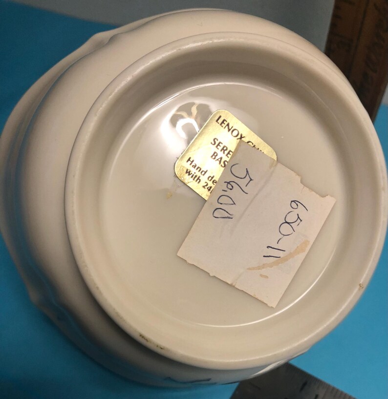 May include: Off-white ceramic bowl with a scalloped rim. The bottom of the bowl has a gold sticker that reads "LENOX SERENE BAS" and a handwritten price tag. The bowl is hand decorated with 24K gold.