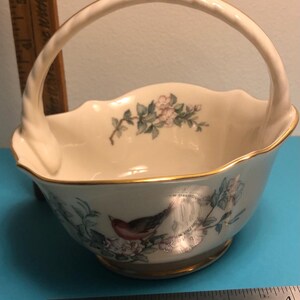 May include: Cream-colored ceramic basket with a handle, featuring a gold rim and floral and bird illustrations. The basket has a scalloped edge and a round base. A ruler is next to the basket.