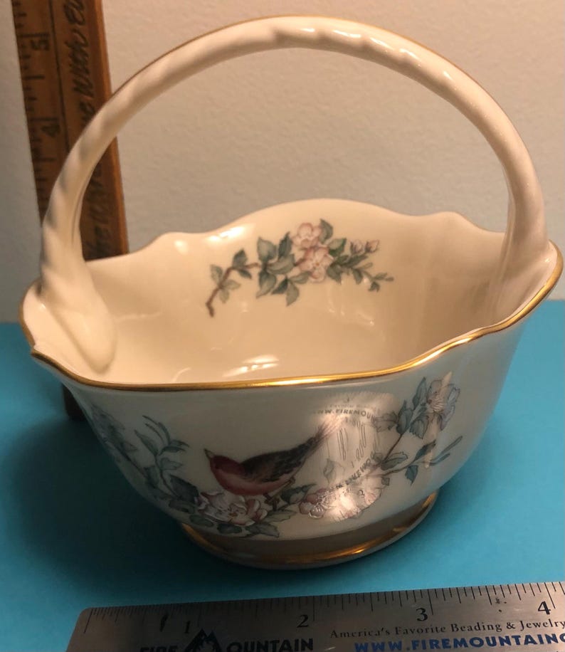 May include: Cream-colored ceramic basket with a handle, featuring a gold rim and floral and bird illustrations. The basket has a scalloped edge and a round base. A ruler is next to the basket.