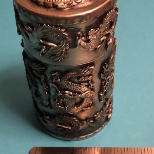 Può includere: Un contenitore cilindrico in metallo con intricate sculture a rilievo di draghi. Il coperchio presenta un design dettagliato di drago. Il contenitore è alto circa 5 cm e ha una finitura metallica. Tema di ispirazione asiatica.