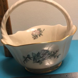 May include: Cream-colored ceramic basket with a handle and gold trim. The basket features floral designs in pink and green. The handle is twisted, and the rim has a scalloped edge. A decorative piece for the home.