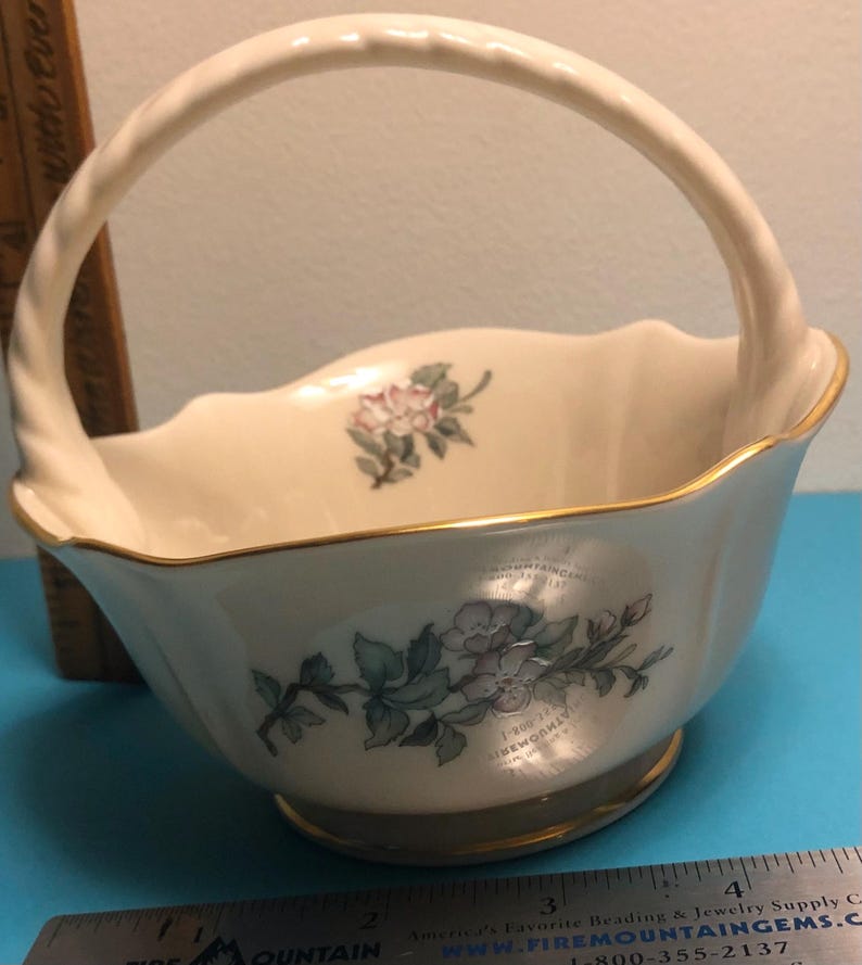 May include: Cream-colored ceramic basket with a handle and gold trim. The basket features floral designs in pink and green. The handle is twisted, and the rim has a scalloped edge. A decorative piece for the home.