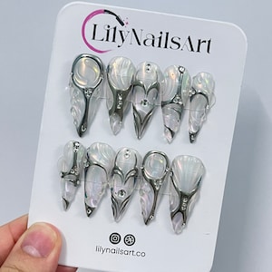 May include: Set of ten white and silver press-on nails with a pearl-like finish. Each nail features a different design with silver accents and rhinestones. The nails are displayed on a white card with the text "LilyNailsArt" and "lilynailsart.co".