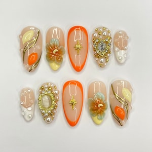 May include: Eight peach-colored nail tips with various designs. The designs include a flower, a star, a heart, and a swirl pattern. The nail tips are decorated with gold and white accents.