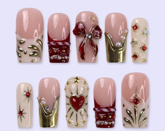 May include: A set of ten press-on nails featuring a nude base with various decorative elements including gold metallic accents, red rhinestones, white pearls, a red bow, a red heart, and star-shaped embellishments in a glossy finish.