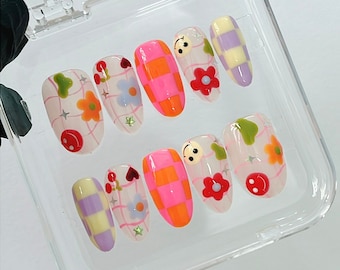 May include: A set of ten colorful press-on nails with various designs, including flowers, smiley faces, and checkered patterns. The nails are displayed in a clear plastic case.