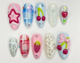 May include: Ten different nail designs featuring various patterns and colors, including a star, plaid, cherries, a bow, a heart, a strawberry, and swirls. The designs are painted on a white background.