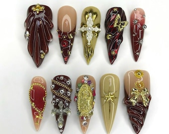 Puede incluir: Conjunto de diez uñas postizas largas y puntiagudas con varios diseños religiosos y ornamentados. Las uñas están decoradas con acentos dorados, plateados y rojos, y presentan cruces, una Virgen María y otros símbolos religiosos.