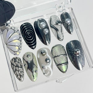 May include: A clear plastic box with ten different nail designs. The designs include swirls, abstract shapes, and glitter. The designs are in shades of white, black, and silver.