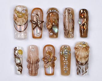 May include: A collection of ten artificial nails with various designs. The nails feature a range of embellishments, including seashells, pearls, and gold accents. The color palette includes shades of brown, beige, and gold, creating a luxurious and elegant aesthetic.