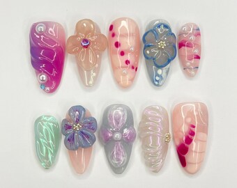 May include: Ten different nail designs featuring a variety of colors and patterns, including pink, blue, green, and white. Some nails have flowers, while others have swirls or dots.