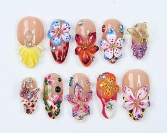 May include: A collection of ten artificial nails, each adorned with intricate floral designs and embellishments. The nails feature a variety of colors, including pink, blue, orange, and yellow, with sparkling rhinestones. The nail shapes are oval and almond.