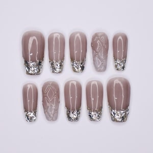 Puede incluir: Diez uñas postizas de color nude con puntas de purpurina plateada. Las uñas tienen una variedad de diseños, que incluyen un patrón geométrico y un diseño de purpurina simple.